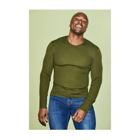 Bald Black man wearing an olive green long-sleeve sweater and blue jeans against a pale green seamless backdrop.