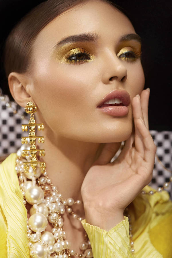 Close-up shot of a woman with golden eyeshadow wearing gold jewelry, against a black and white checkered background.