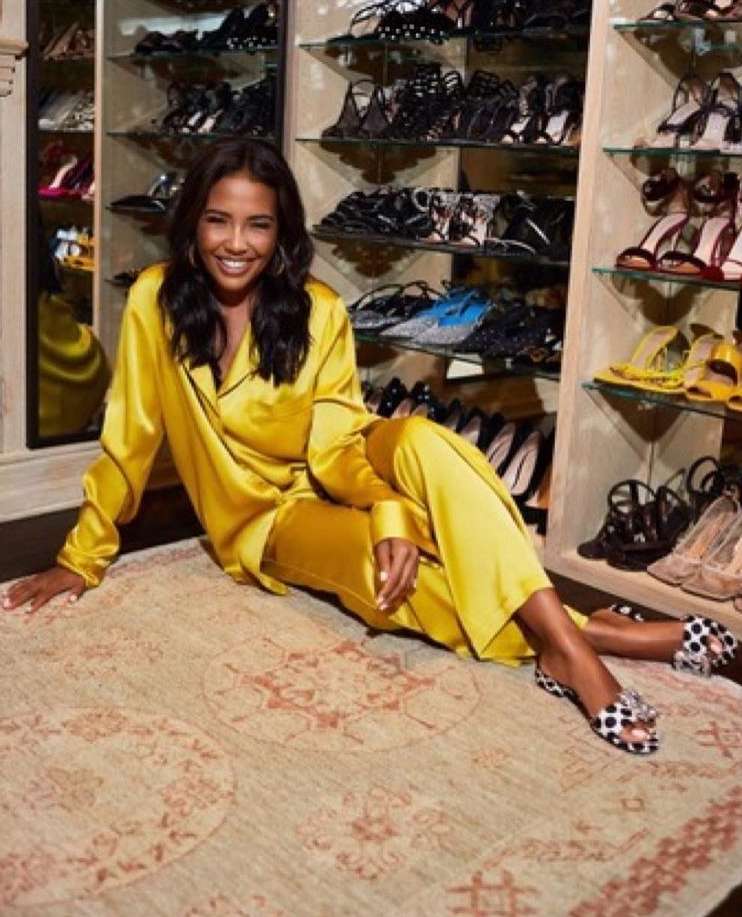 Adult woman in a yellow satin pajama set is sitting on a patterned rug in front of a closet full of shoes and sandals.