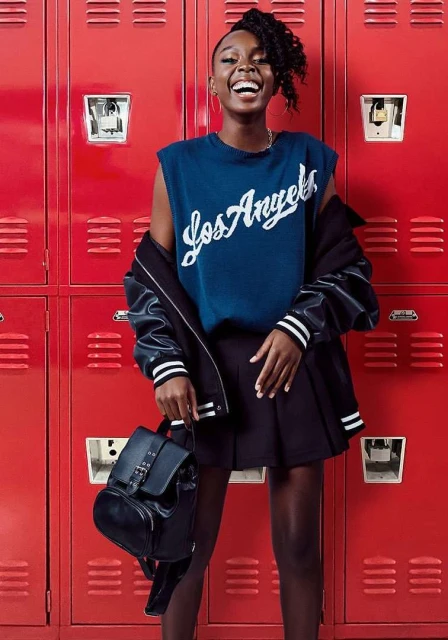 A happy African-American woman stands in front of red lockers wearing a blue shirt, black skirt, and black jacket with a matching backpack.