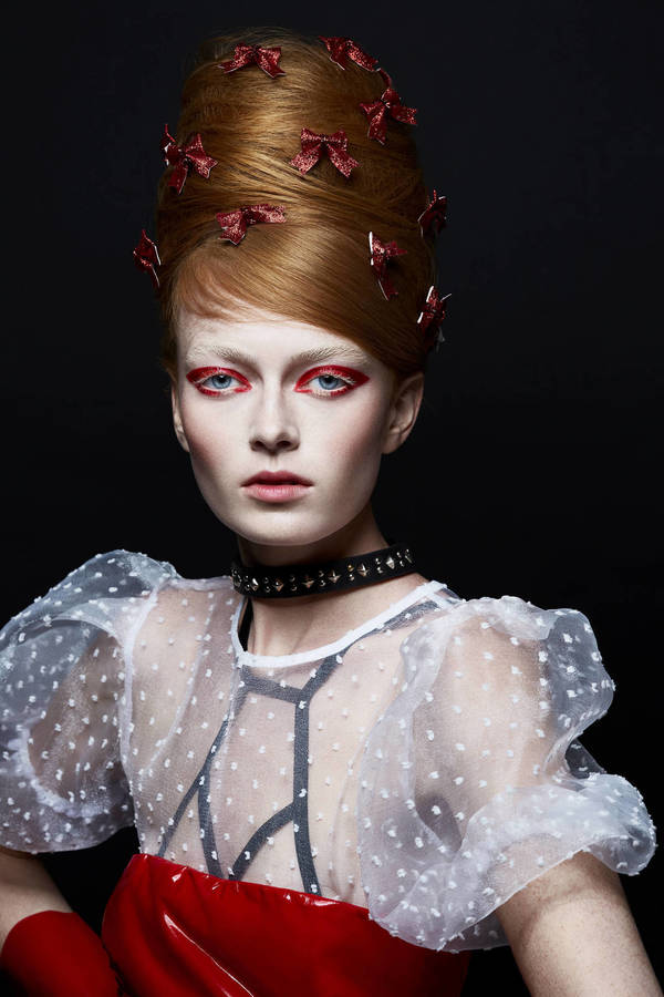 Portrait of a redhead woman with a beehive hairstyle adorned with bows and dramatic red eye makeup, wearing a sheer polka dot top.