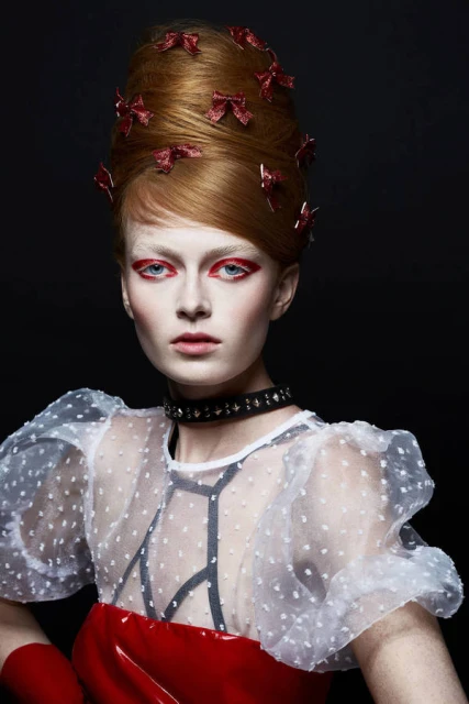 Portrait of a redhead woman with a beehive hairstyle adorned with bows and dramatic red eye makeup, wearing a sheer polka dot top.