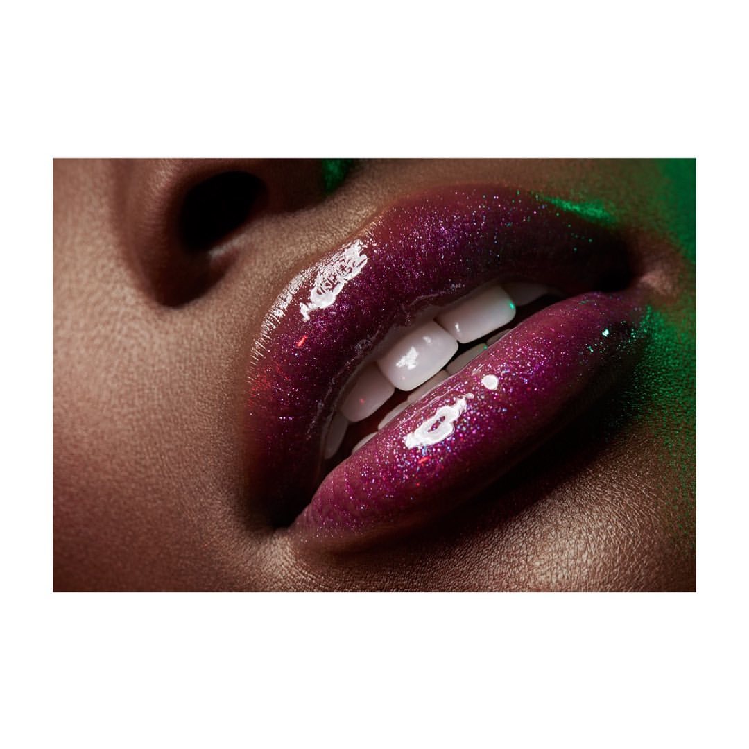Close-up studio shot featuring a woman's open mouth and teeth with glossy purple-magenta lipstick on her lips, with a green background.