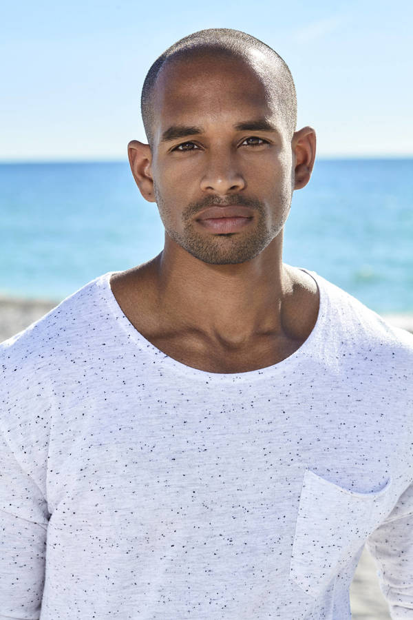 Portrait of a black man wearing a white speckled long-sleeve t-shirt against an ocean beach background.