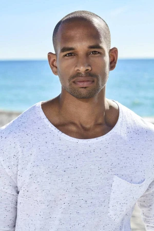 Portrait of a black man wearing a white speckled long-sleeve t-shirt against an ocean beach background.