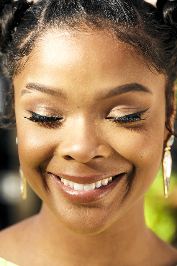 Close-up of a smiling African-American woman with braided space buns, wearing makeup and jewelry. She has her eyes closed.