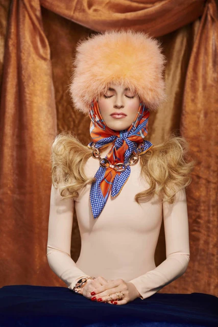 Woman with closed eyes wears a peach hat, scarf, and dress against a tan drapery backdrop, creating a surreal fashion portrait.