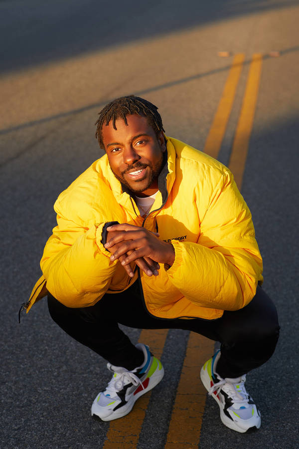 An African-American man in a yellow puffer jacket squats on a road, smiling into the camera with colorful sneakers and black pants.
