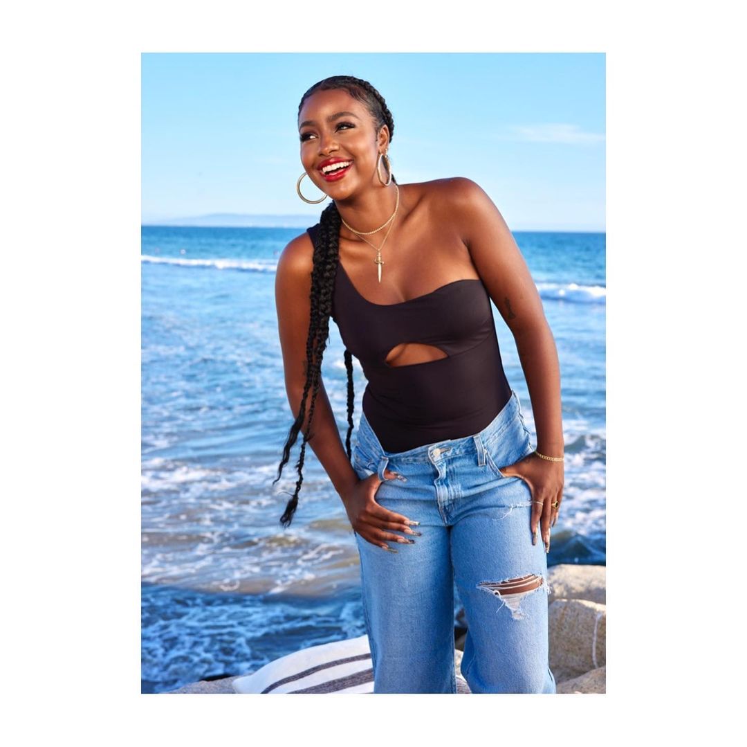 A smiling woman with long braids in a one-shoulder black top and blue jeans stands on a beach with blue sky and ocean backdrop.