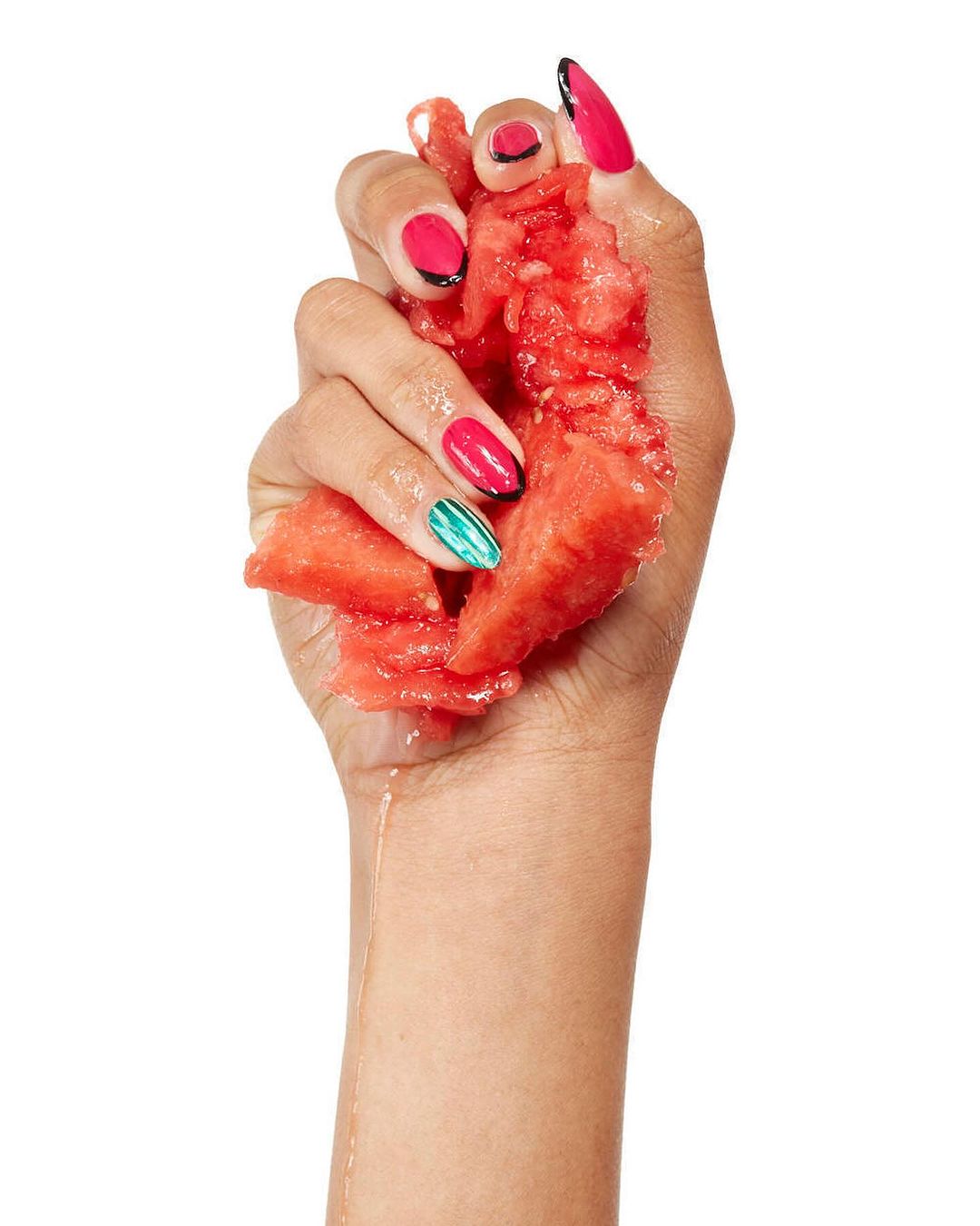 Close-up of a hand squeezing a watermelon, showcasing colorful nail art in pink, red, green, and black against a white background.