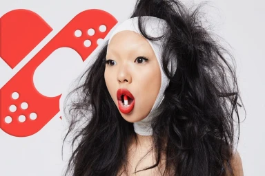 Close-up portrait of a woman with wild black hair, white head bandage, and red lipstick holding a capsule pill in her mouth.