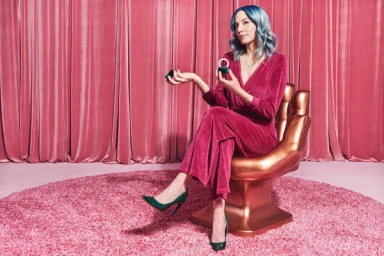 Woman with blue hair wearing a magenta jumpsuit seated on a bronze hand-shaped chair holding a small box