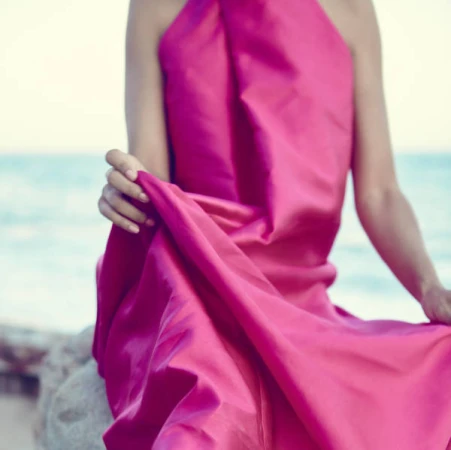Woman wearing a bright pink halter dress sitting by the ocean holding the hem of the garment