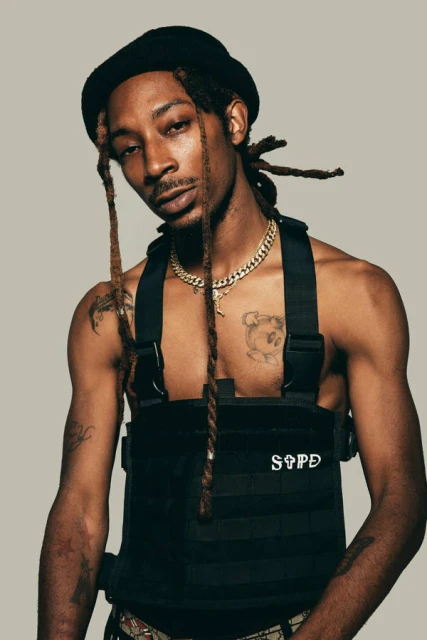 Portrait of a Black male model wearing a black vest and a gold chain with dreadlocks and a black beanie against a grey background.