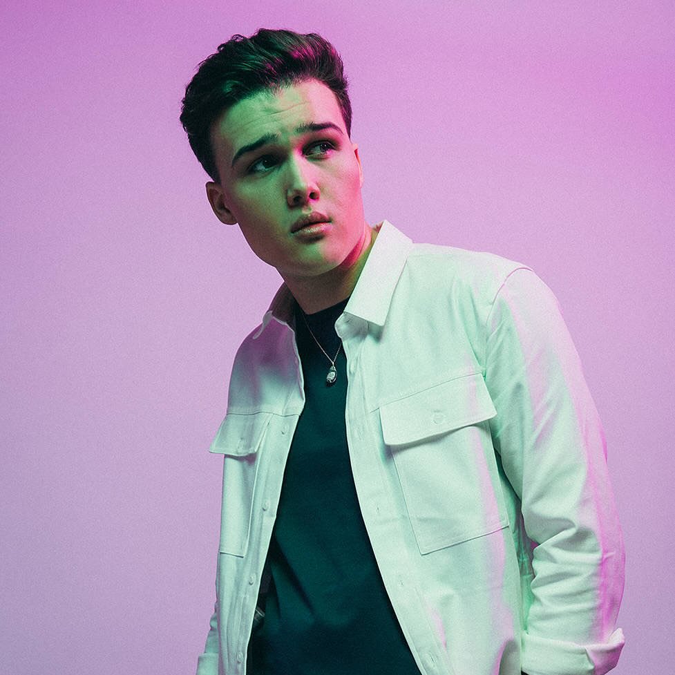 Portrait of a young adult man wearing a white jacket, looking off to the side in a photo studio with soft pink and green lighting.