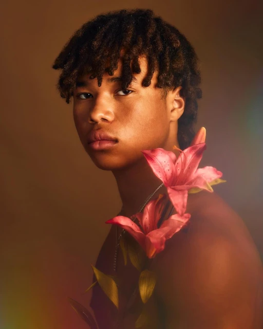 Close-up of a young adult with dark hair, posing with pink lilies against a warm brown background in a portrait orientation.