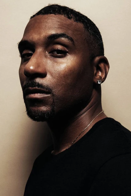 Portrait of an adult Black male in a dark T-shirt against a tan background. The subject is facing slightly to the left with a neutral facial expression.