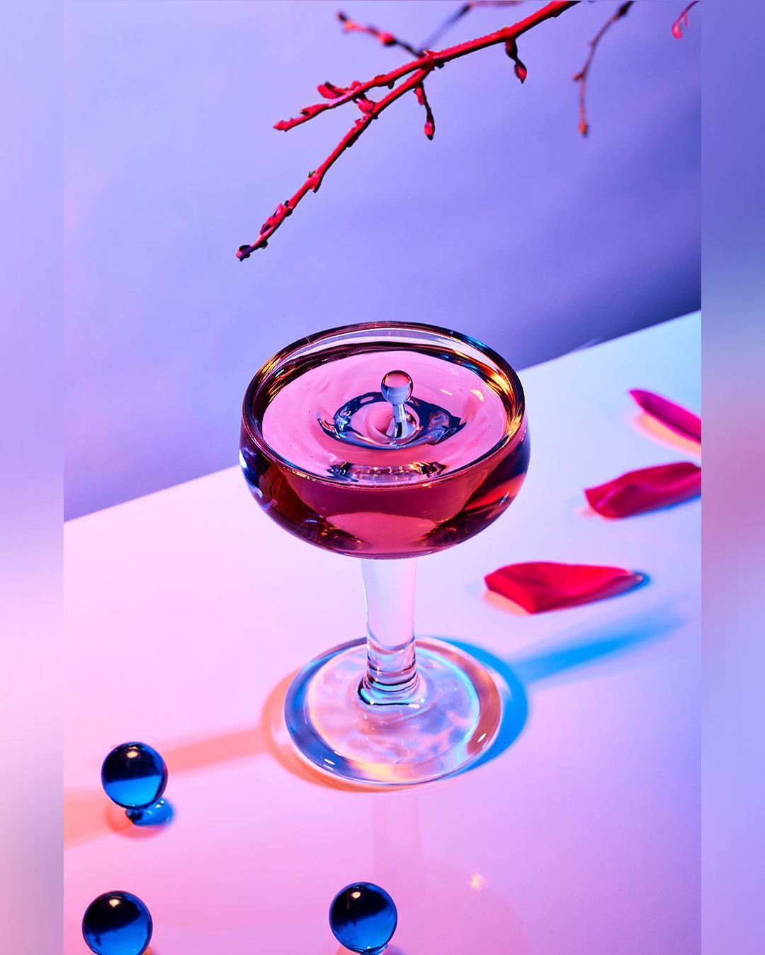 Close-up of a wine glass with liquid and a water drop, alongside scattered rose petals and blue glass marbles on a white surface.