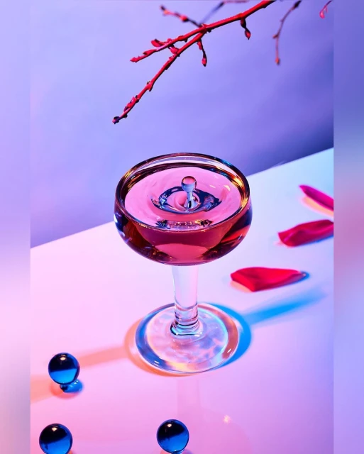 Close-up of a wine glass with liquid and a water drop, alongside scattered rose petals and blue glass marbles on a white surface.