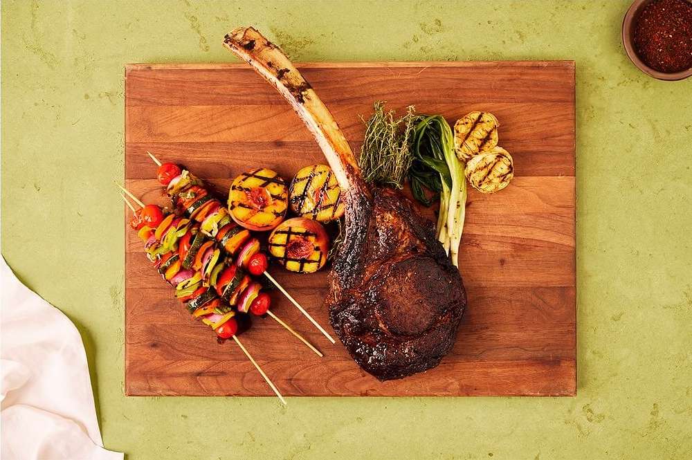 Elevated shot of grilled steak, vegetable skewers, and peaches on a wooden board set against a light green backdrop, gourmet food.