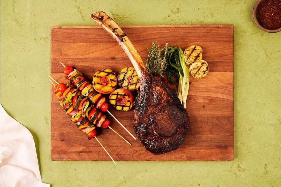 Elevated shot of grilled steak, vegetable skewers, and peaches on a wooden board set against a light green backdrop, gourmet food.