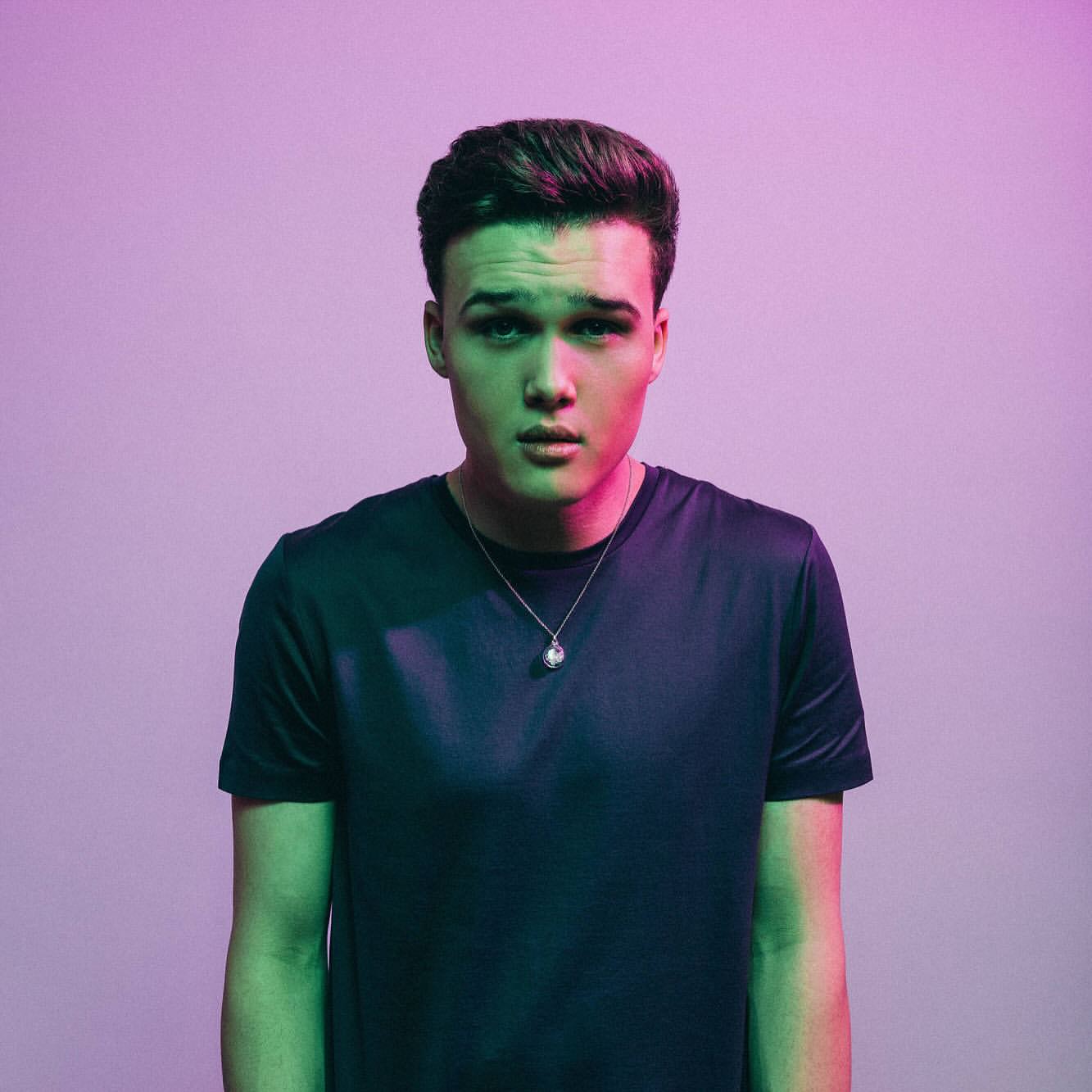 Portrait of a young adult male in a dark t-shirt looking directly at the camera, lit with green and purple gel lighting.