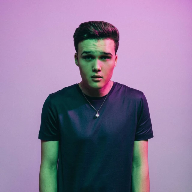 Portrait of a young adult male in a dark t-shirt looking directly at the camera, lit with green and purple gel lighting.
