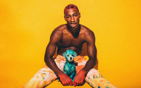 Shirtless man with red hair sits with a blue-dyed puppy against a yellow backdrop, wearing patterned tie-dye pants, in a vibrant studio portrait.