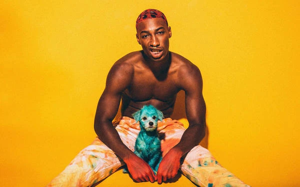 Shirtless man with red hair sits with a blue-dyed puppy against a yellow backdrop, wearing patterned tie-dye pants, in a vibrant studio portrait.