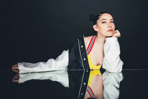 Woman with hair in double buns leaning on a reflective surface wearing a black and white jacket over a yellow top