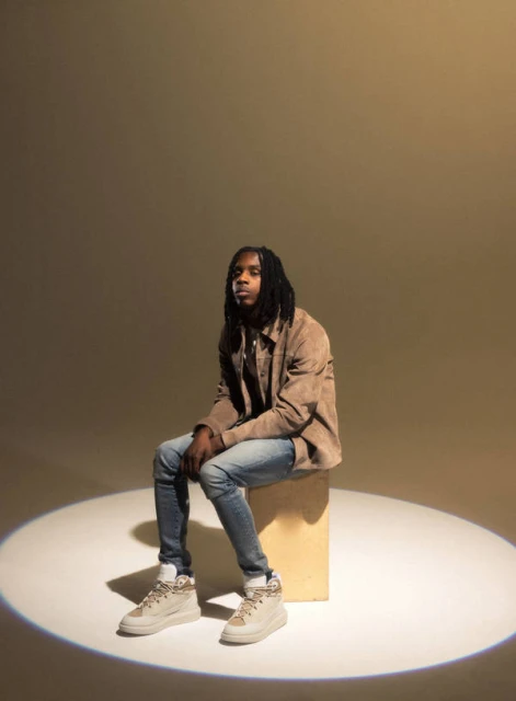Adult male with dreadlocks wearing a tan jacket and jeans sitting on a wooden cube in a photo studio with spotlight lighting.