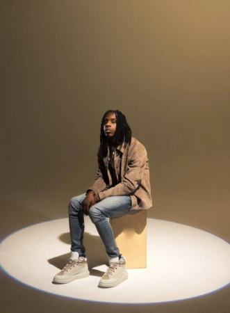 Adult male with dreadlocks wearing a tan jacket and jeans sitting on a wooden cube in a photo studio with spotlight lighting.