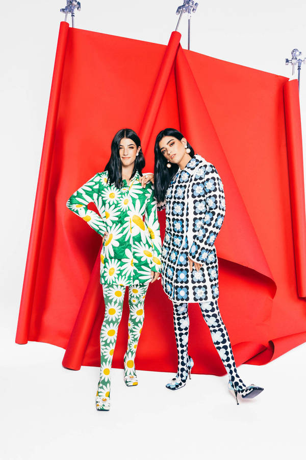 Two women posing against a red paper backdrop, wearing matching patterned dresses and patterned tights.