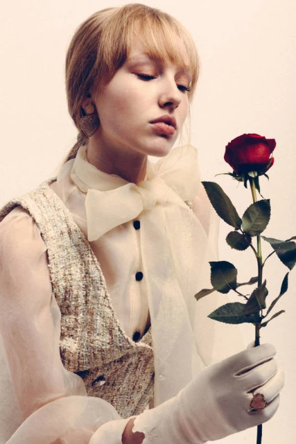 A young woman with blonde hair in a sheer blouse holds a red rose against a neutral background in a fashion forward portrait.