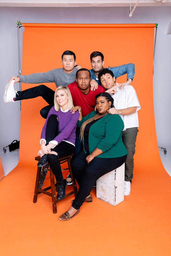 Group photo of six adults in casual clothing in a photo studio with an orange backdrop, posing and smiling at the camera.
