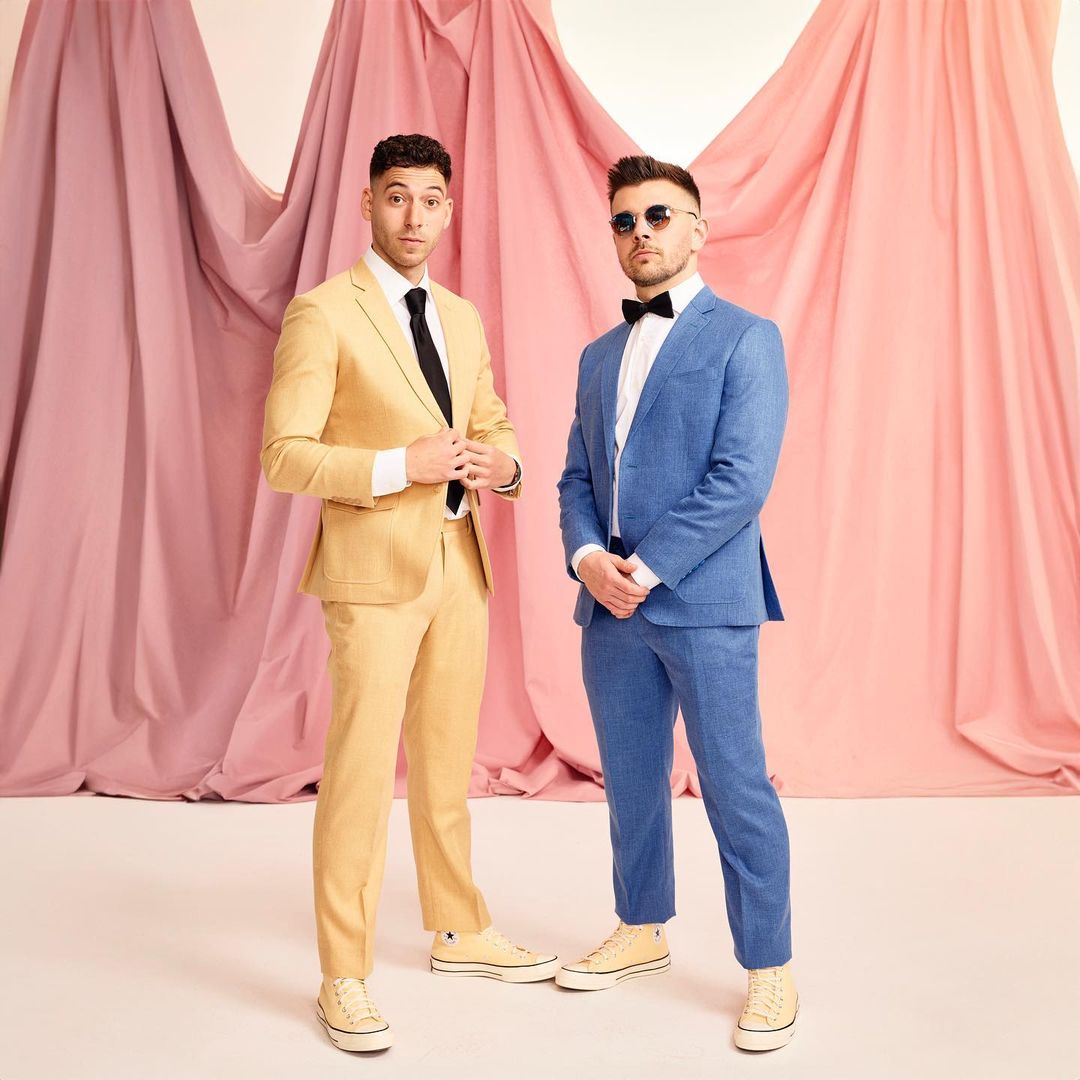 Two men in suits stand in front of a backdrop of pink and white draped fabric. One wears a yellow suit, the other a blue suit.