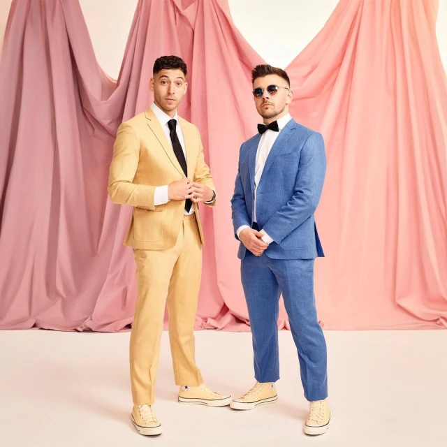 Two men in suits stand in front of a backdrop of pink and white draped fabric. One wears a yellow suit, the other a blue suit.