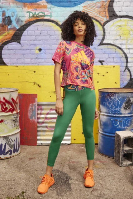 Woman wearing a colorful patterned t-shirt, green leggings, and orange sneakers, posing against a graffiti wall in an urban setting.