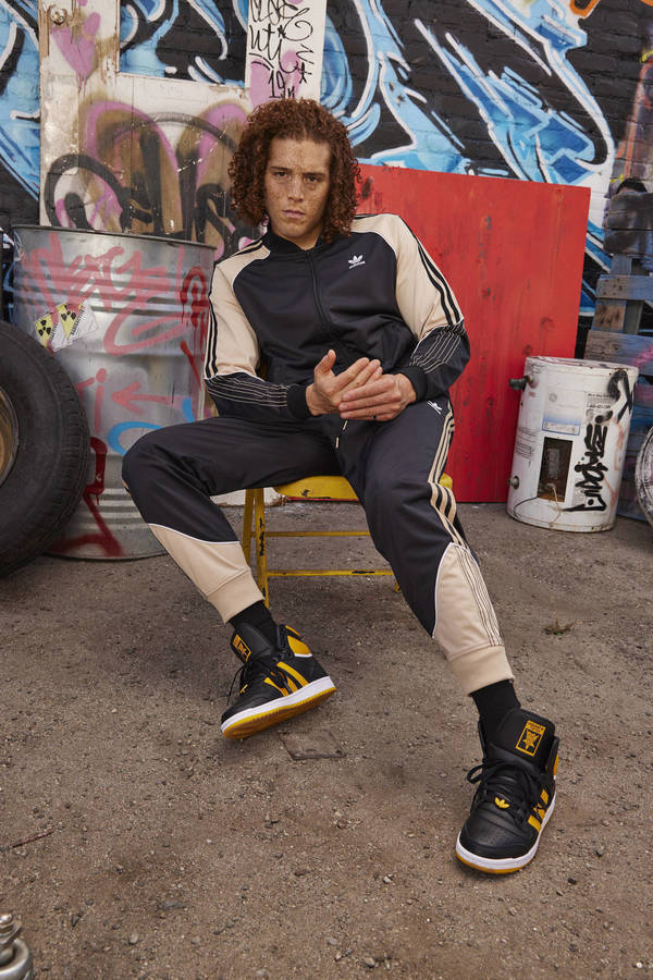 Young man with curly hair wearing a black and beige tracksuit seated on a yellow stool against a graffiti wall