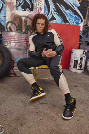 Young man with curly hair wearing a black and beige tracksuit seated on a yellow stool against a graffiti wall