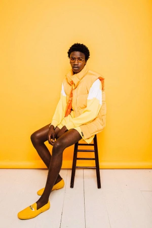 Stylish African American man in orange vest and loafers sitting on stool against bright yellow backdrop in studio environment. 