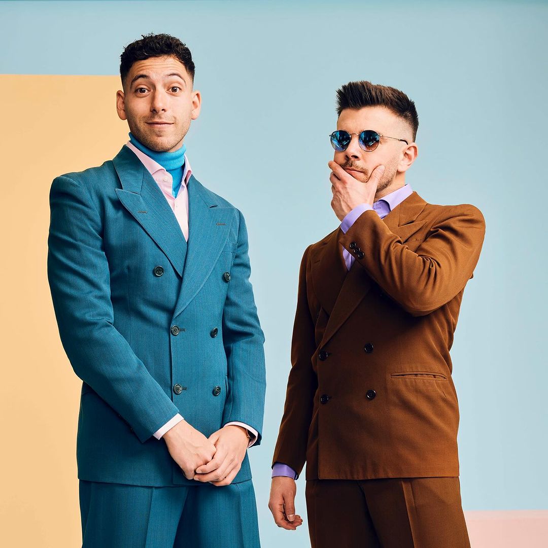 Two men in suits stand side-by-side against a colorful geometric backdrop, one in brown, one in teal, in a studio setting with good lighting.