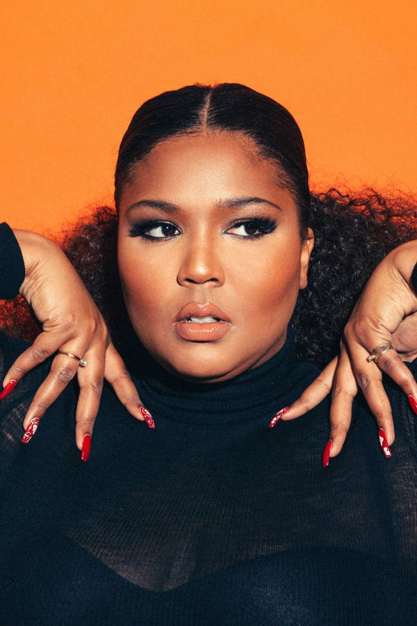 Portrait of a woman with an orange backdrop wearing a black shirt with long red nails and gold rings looking slightly upward.