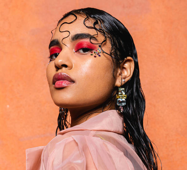 Portrait of an adult female model with red eyeshadow, glittery stars near her eye, a sheer pink top, and an orange textured background.