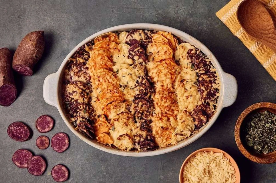 Overhead shot of scalloped purple and orange sweet potatoes, baked in a ceramic dish, alongside spices and a yellow tea towel.