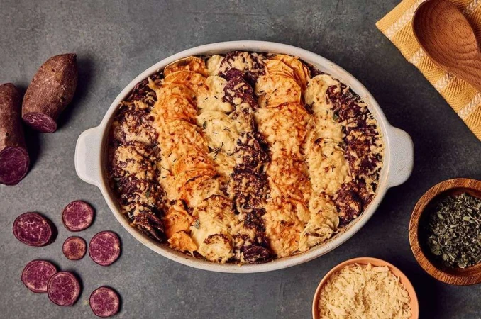 Overhead shot of scalloped purple and orange sweet potatoes, baked in a ceramic dish, alongside spices and a yellow tea towel.