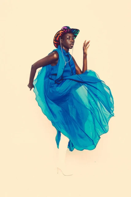 An African woman poses in a bright turquoise chiffon dress with a colorful hat against a studio backdrop, fashion editorial style.