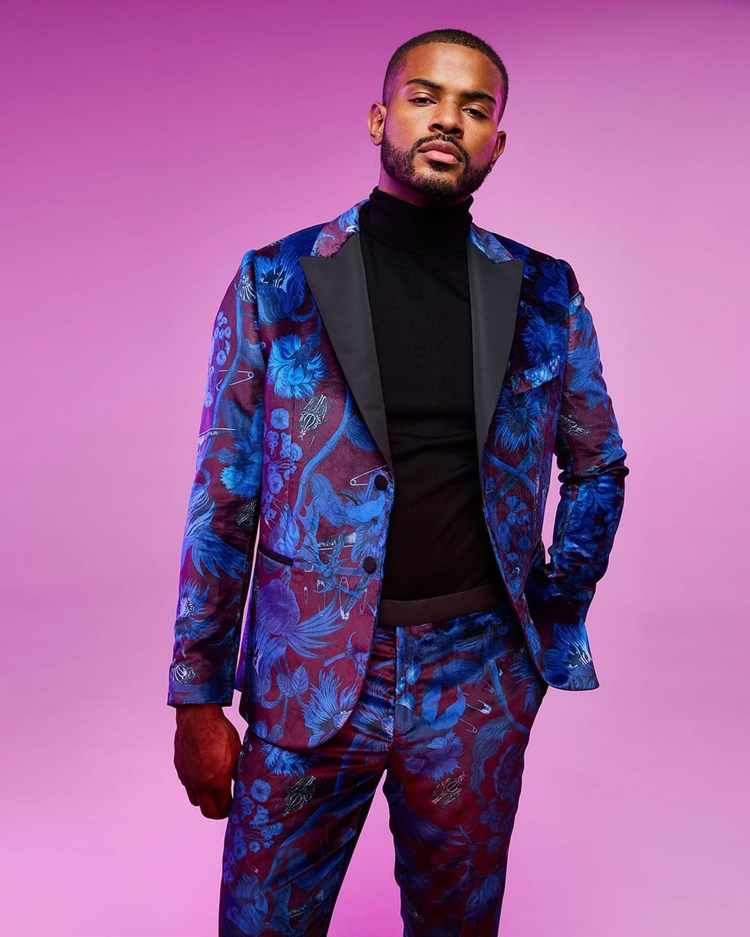 A man poses in a patterned blue and burgundy suit against a purple backdrop in a portrait showcasing fashion and style, with a confident expression.