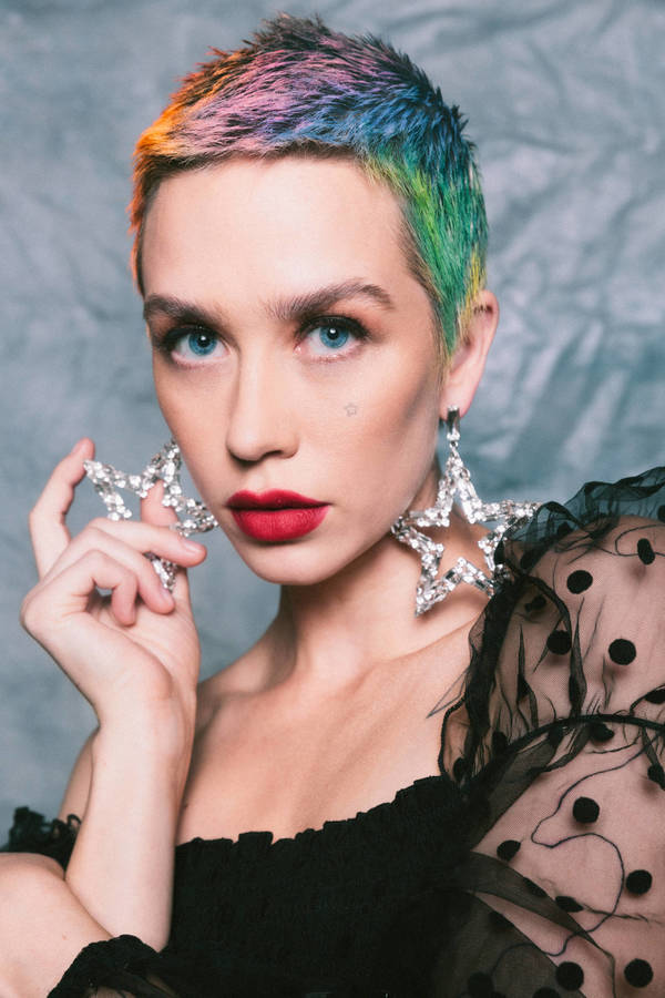 Close-up portrait of a fair-skinned adult female model with rainbow-colored pixie hair, wearing star-shaped earrings and a black dress.