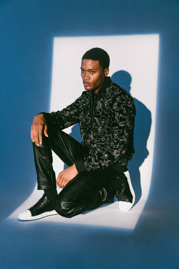 A young Black male model in a black patterned hoodie and shiny black pants sits on a blue background with a white spotlight.